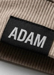 Close-up of a ribbed knit garment in a light taupe-beige color, featuring a black fabric brand label with the name 'ADAM' in white embroidered letters.
